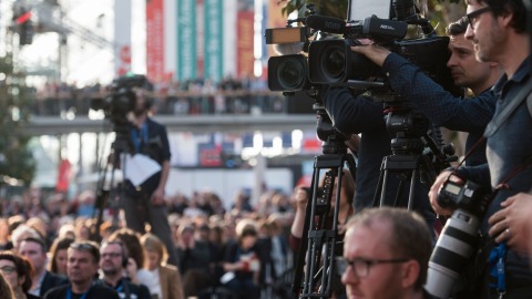 A photo from an event at the Leipzig Book Fair. Three cameramen and their cameras can be seen pointing at an object outside the frame. Two of the men are clearly visible on the right side of the image. One is blurred in the background. 