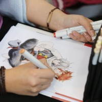 Close-up of a drawing of two manga characters being finished, with pens visible on the right.
