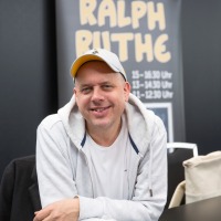 Ralph Rute sits at a signing table at the Manga-Comic-Con 2024, smiling at the camera
