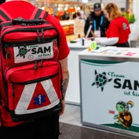 A person from the Awareness Team SAM at the Manga-Comic-Con 2025 walks past a booth, clearly showing the team’s red backpack.
