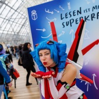 Das Maskottchen der Manga Comic Con Macoco macht einem Luftkuss vor einer bunten Leinwand auf der Manga Comic Con 2025