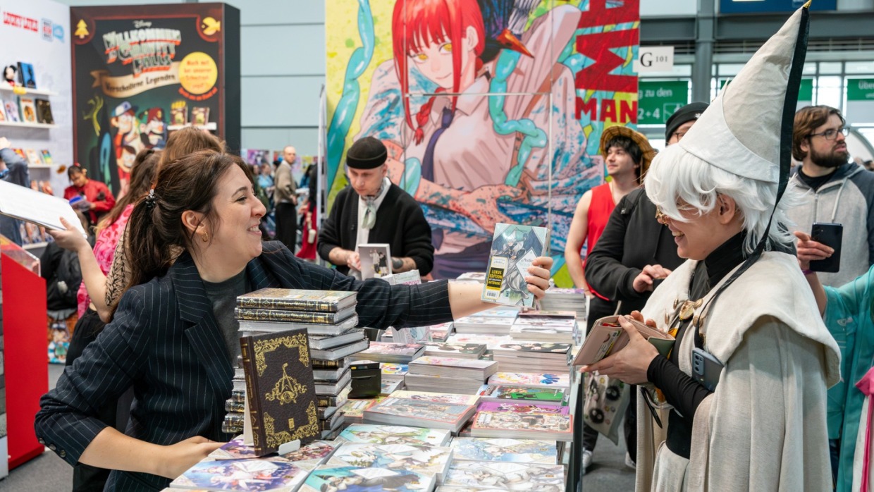 Exhibitor presents cosplayer with a manga comic book from a table full of manga comics at Manga Comic Con 2025.