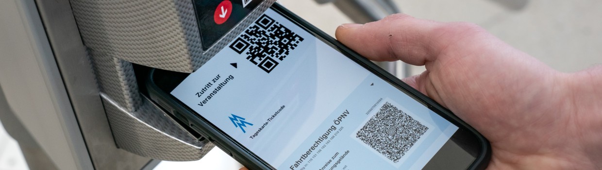 A hand is holding a smartphone under the scanner at the entrance turnstile. The display shows an electronic ticket for the Leipzig Trade Fair with a QR code, including a public transport ticket.

