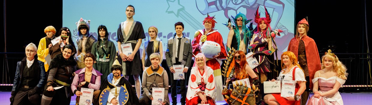 Foto vom Leipziger Cosplay Wettbewerb 2023. Es sind viele verschiedene Cosplayer für ein Abschlussfoto auf der Bühne zu sehen, mit dem Maskottchen MaCoCo im Hinterrgund auf einer Leinwand.