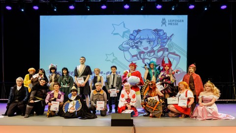 Foto vom Leipziger Cosplay Wettbewerb 2023. Es sind viele verschiedene Cosplayer für ein Abschlussfoto auf der Bühne zu sehen, mit dem Maskottchen MaCoCo im Hinterrgund auf einer Leinwand.
